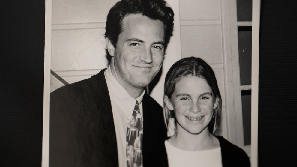 "A black-and-white photo showing a man in a suit and tie smiling next to a young girl with braces. They are indoors, in front of a white wall with window panes. The atmosphere appears warm and friendly."