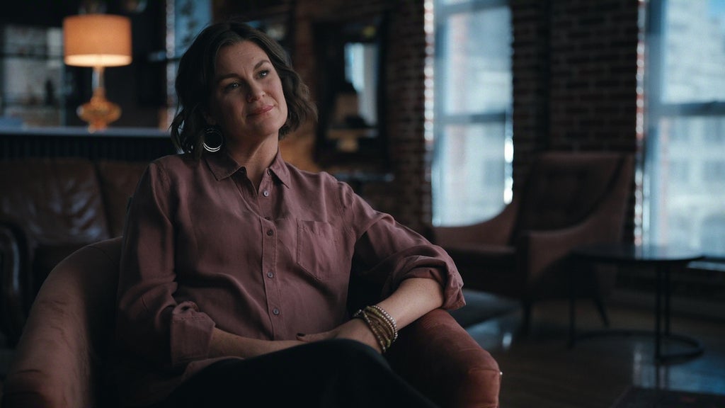 "Woman sitting in a modern, cozy loft with brick walls and large windows, soft lighting, armchairs, and a lamp in the background, looking relaxed and thoughtful."