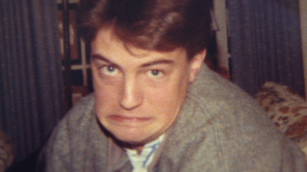 "Young man with brown hair making a humorous, exaggerated facial expression indoors. He is wearing a grey jacket. The background features curtains and furniture, suggesting a casual home setting."
