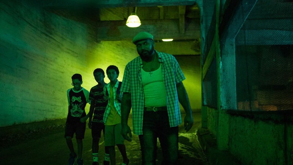 Four figures walk toward the camera through a dim concrete tunnel bathed in eerie green light, with a hanging lamp overhead.